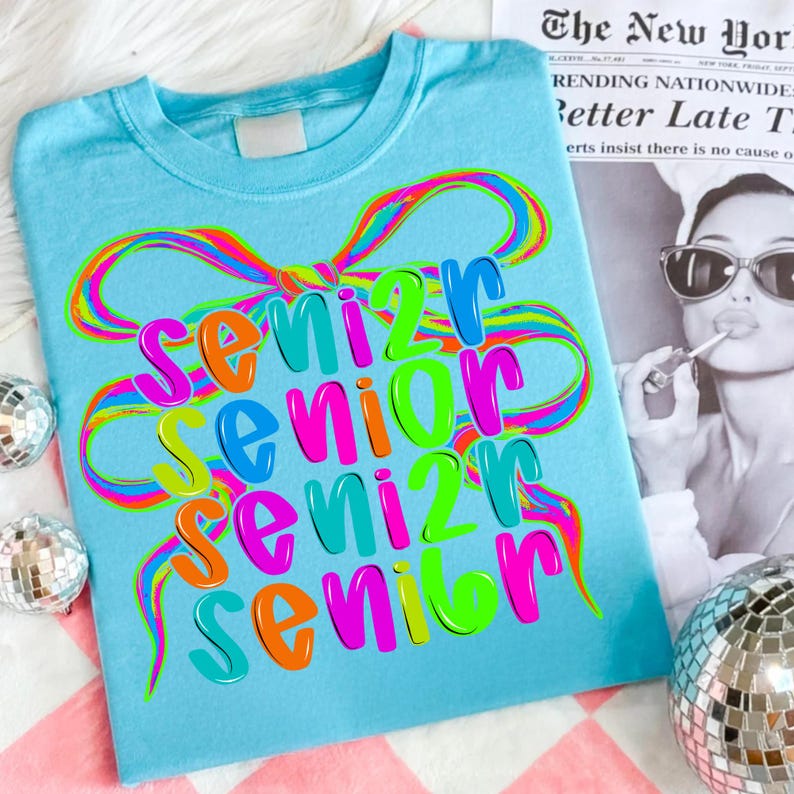 May include: A light blue t-shirt with the word "Senior" in a colorful, layered design. The word is repeated four times, with a rainbow-colored bow graphic above. The shirt is on a pink and white checkered surface.
