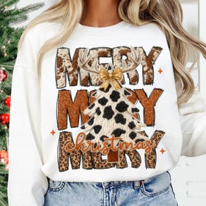 May include: White sweatshirt with a festive "Merry Christmas" design. The design features the words "Merry Christmas" in a mix of brown, leopard print, and cow print patterns, with a Christmas tree and antlers.