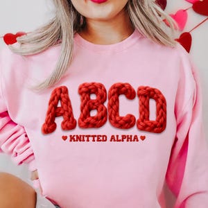 May include: A light pink sweatshirt with the letters ABCD in a chunky, red, knitted style. Below the letters, the text reads "KNITTED ALPHA" with heart symbols. The sweatshirt is a solid color.