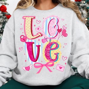 以下が含まれることがあります： カラフルで気まぐれなデザインの「LOVE」という文字がプリントされたライトグレーのスウェットシャツ。文字はハート、リボン、その他のバレンタインデーをテーマにしたグラフィックで装飾されています。「L」は黄色とピンク、「O」は青とピンク、「V」はピンク、「E」は黄色とピンクです。