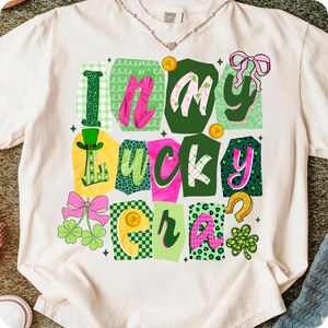 May include: Cream-colored t-shirt with the phrase "In My Lucky Era" in a colorful, patchwork design. The design includes green, pink, and yellow elements, such as shamrocks, a horseshoe, and a leprechaun hat, perfect for St. Patrick's Day.