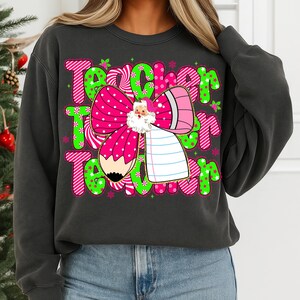 Peut inclure: Sweat-shirt gris fonc&eacute; avec un motif graphique pour les enseignants. Le motif pr&eacute;sente le mot "Teacher" en rose et vert, un n&oelig;ud rose &agrave; pois blancs, une illustration du P&egrave;re No&euml;l, un crayon et un bloc-notes.