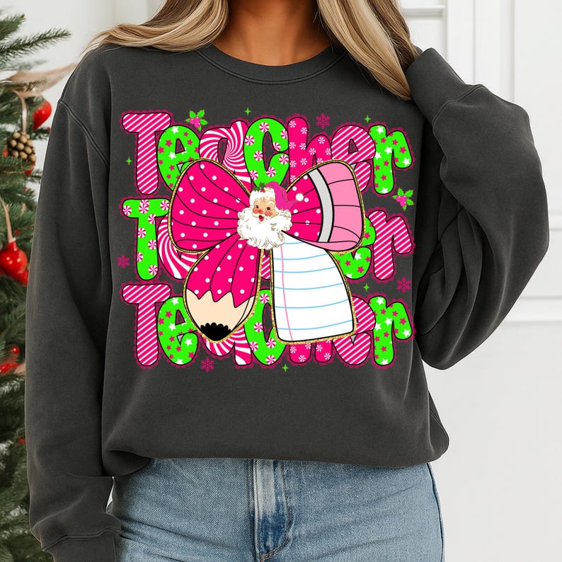 Peut inclure: Sweat-shirt gris fonc&eacute; avec un motif graphique pour les enseignants. Le motif pr&eacute;sente le mot "Teacher" en rose et vert, un n&oelig;ud rose &agrave; pois blancs, une illustration du P&egrave;re No&euml;l, un crayon et un bloc-notes.