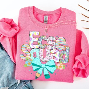 May include: A pink sweatshirt with the text "Ecse Squad" in a colorful, patterned design. The design includes a turquoise bow, a pencil, and floral accents. A silver necklace with a pendant is also visible.