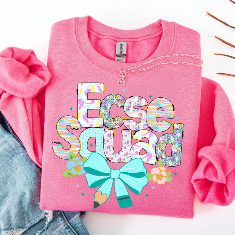 May include: A pink sweatshirt with the text "Ecse Squad" in a colorful, patterned design. The design includes a turquoise bow, a pencil, and floral accents. A silver necklace with a pendant is also visible.