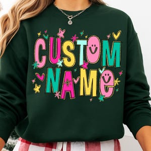 May include: Dark green sweatshirt with the words "CUSTOM NAME" in colorful, playful lettering. The text is outlined in gold and decorated with stars, hearts, and smiley faces. A fun, personalized apparel item.