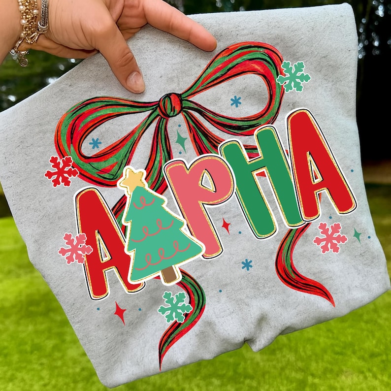 May include: A light gray sweatshirt featuring the word "ALPHA" in red, pink, and green, with a Christmas tree graphic. The design includes a large bow, snowflakes, and stars in red, green, and blue.