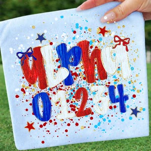 May include: Light blue fabric with the word "ALPHA" in red, white, and blue letters, and the numbers "01234". The design includes red and blue paint splatters, gold glitter, and star and bow accents. The fabric is held by a hand.