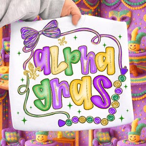 May include: White sweatshirt with the text "Alpha Gras" in purple, green, and yellow, with gold glitter accents. The design includes a purple bow, fleur-de-lis, and a strand of beads. The sweatshirt is held up by a person.