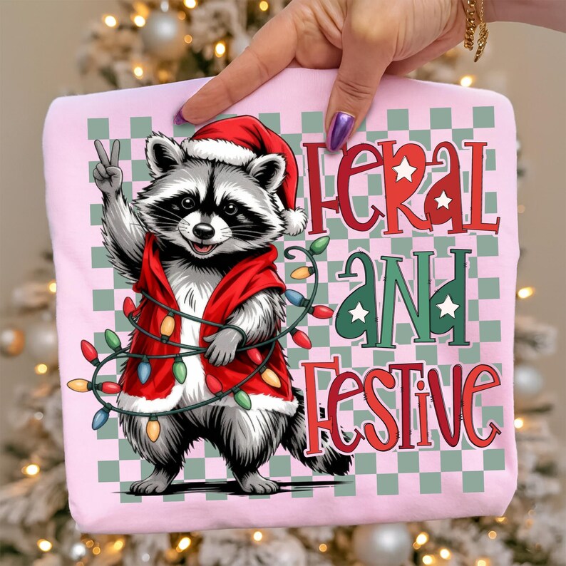 May include: A light pink t-shirt featuring a raccoon wearing a Santa hat and a red vest, holding a string of Christmas lights and giving a peace sign. The text "Feral and Festive" is printed on the shirt.