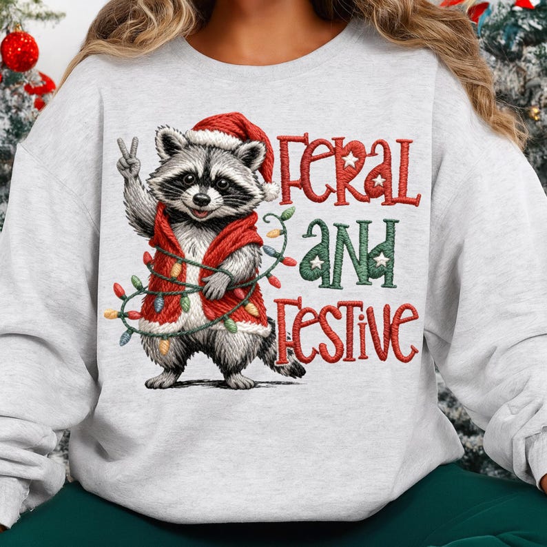 May include: A light gray sweatshirt featuring a raccoon wearing a Santa hat and a red vest adorned with Christmas lights. The text "Feral and Festive" is embroidered in red and green. The raccoon is giving a peace sign.