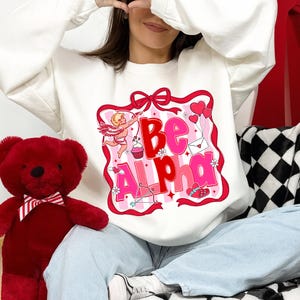 May include: White sweatshirt with a pink and red graphic that says "Be Alpha". The graphic includes a cupid, hearts, and a bow. A red teddy bear with a bow tie is on the left.