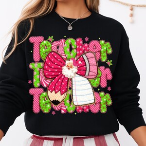 May include: Black sweatshirt with a colorful graphic design. The design features the word "Teacher" in pink and green, a pink bow, a pencil, and a Santa Claus figure. The sweatshirt is a festive holiday item.