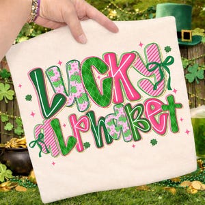 Pu&ograve; includere: Una federa color crema con la scritta "Lucky Alphabet" in un design colorato. Le lettere sono bordate di verde e rosa, con motivi a trifoglio e a righe. Un fiocco verde adorna la lettera "J".