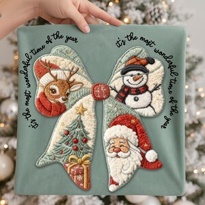 May include: A decorative Christmas pillow with a bow design featuring a reindeer, snowman, Christmas tree, and Santa Claus. The text "It's the most wonderful time of the year" is written around the bow in black script. The pillow is held by a hand.