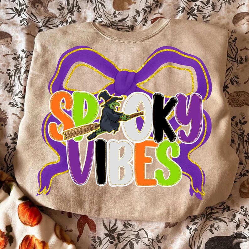 May include: Beige crewneck sweatshirt with the words "Spooky Vibes" in orange, green, black, and purple. A witch on a broom is in the center. The design includes a purple bow with gold glitter accents. A Halloween-themed sweatshirt.