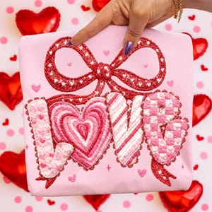May include: A light pink tote bag with a red embroidered bow and the word "LOVE" in a decorative font. The letters are in pink and white patterns, with heart and star accents. The bag is held up against a background of red heart-shaped balloons.