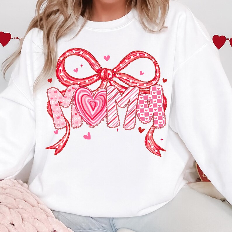 May include: White sweatshirt featuring a large red bow with heart accents and the word "MOM" in a decorative font. The letters have various patterns, including hearts and stripes. Small pink hearts are scattered around the design, creating a festive look.