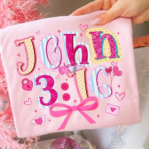 May include: A light pink t-shirt with the text "John 3:16" in a decorative font. The design includes hearts, a bow, and other romantic elements. The shirt is held up by a person.