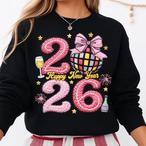 May include: Black sweatshirt with a colorful embroidered design for the New Year. The design includes the numbers "26" in pink, a disco ball, a champagne glass, and the words "Happy New Year". Gold stars and fireworks complete the festive look.