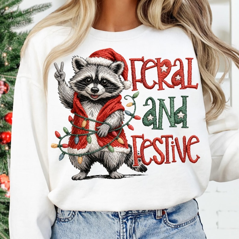 May include: White sweatshirt with a graphic of a raccoon wearing a Santa hat and a red vest, holding a string of Christmas lights. The text "FERAL AND FESTIVE" is in red and green.