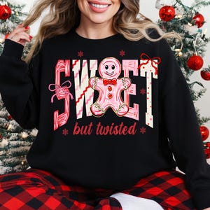May include: Black sweatshirt featuring a pink and white graphic with the text "SWEET" and a gingerbread man, plus the words "but twisted". The design includes snowflakes and bows. The sweatshirt has a crew neck and long sleeves.