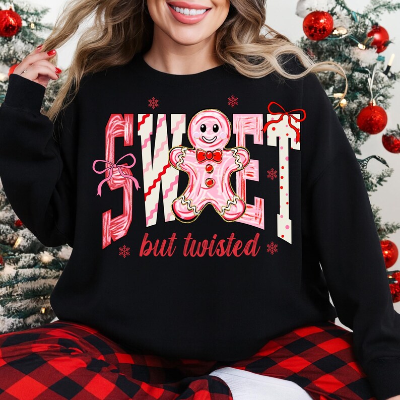 May include: Black sweatshirt featuring a pink and white graphic with the text "SWEET" and a gingerbread man, plus the words "but twisted". The design includes snowflakes and bows. The sweatshirt has a crew neck and long sleeves.