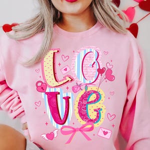 以下が含まれることがあります： ピンク色のスウェットシャツ。カラフルなデザインで「LOVE」の文字が描かれています。文字はハート、バラ、リボンで飾られています。「L」はピンクのドット柄、「O」は青と白のストライプ、「V」はピンクと青のストライプ、「E」はピンクの花柄です。