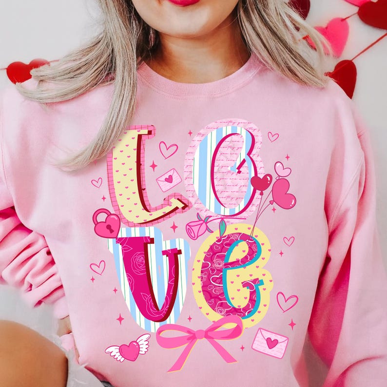 以下が含まれることがあります： ピンク色のスウェットシャツ。カラフルなデザインで「LOVE」の文字が描かれています。文字はハート、バラ、リボンで飾られています。「L」はピンクのドット柄、「O」は青と白のストライプ、「V」はピンクと青のストライプ、「E」はピンクの花柄です。