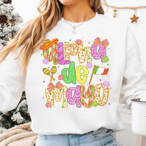 K&ouml;nnte beinhalten: Wei&szlig;es Sweatshirt mit dem Text "Alpha de Mayo" in farbenfrohen, festlichen Buchstaben. Das Design umfasst einen Sombrero, Maracas, Blumen, eine Schleife und die mexikanische Flagge. Ein lustiges, festliches Design.