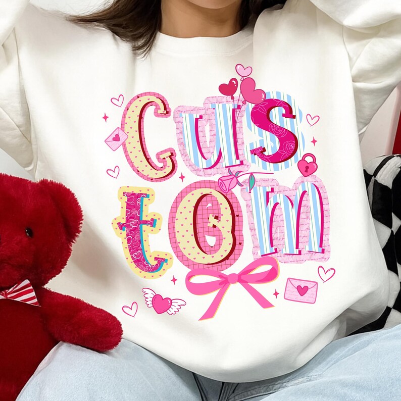 May include: White sweatshirt with the word "Custom" in a colorful, whimsical design. The letters are outlined in pink and decorated with hearts, roses, and a bow. The sweatshirt is a casual, comfortable style.