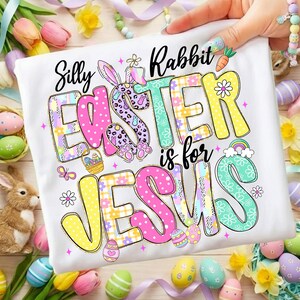 May include: White t-shirt with the text "Silly Rabbit Easter is for Jesus" in colorful, patterned letters. The design includes Easter eggs, a bunny, and a rainbow. The shirt is decorated with floral and polka dot patterns.