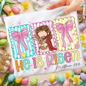 May include: White t-shirt with an Easter design. The design includes Jesus holding a rabbit, pink bows, and the text "He Is Risen" with "Matthew 28:6". The design is surrounded by Easter eggs and flowers.