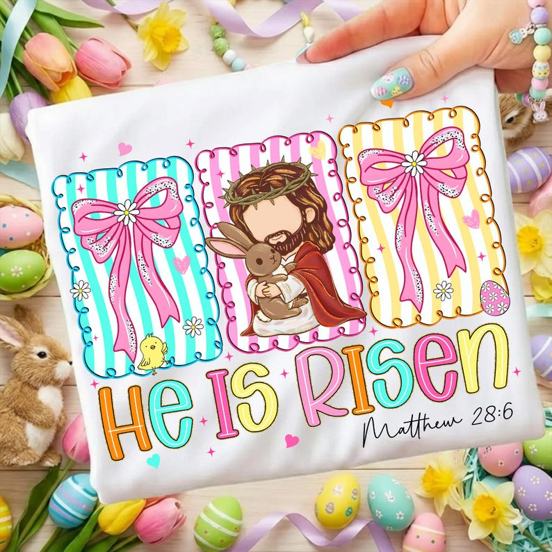 May include: White t-shirt with an Easter design. The design includes Jesus holding a rabbit, pink bows, and the text "He Is Risen" with "Matthew 28:6". The design is surrounded by Easter eggs and flowers.