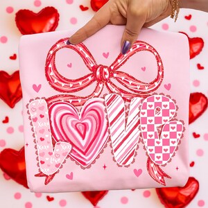 May include: A light pink t-shirt with a large red bow and the word "LOVE" in a decorative font. The "O" is a heart shape. The shirt has small heart and "XOXO" details, perfect for Valentine's Day.