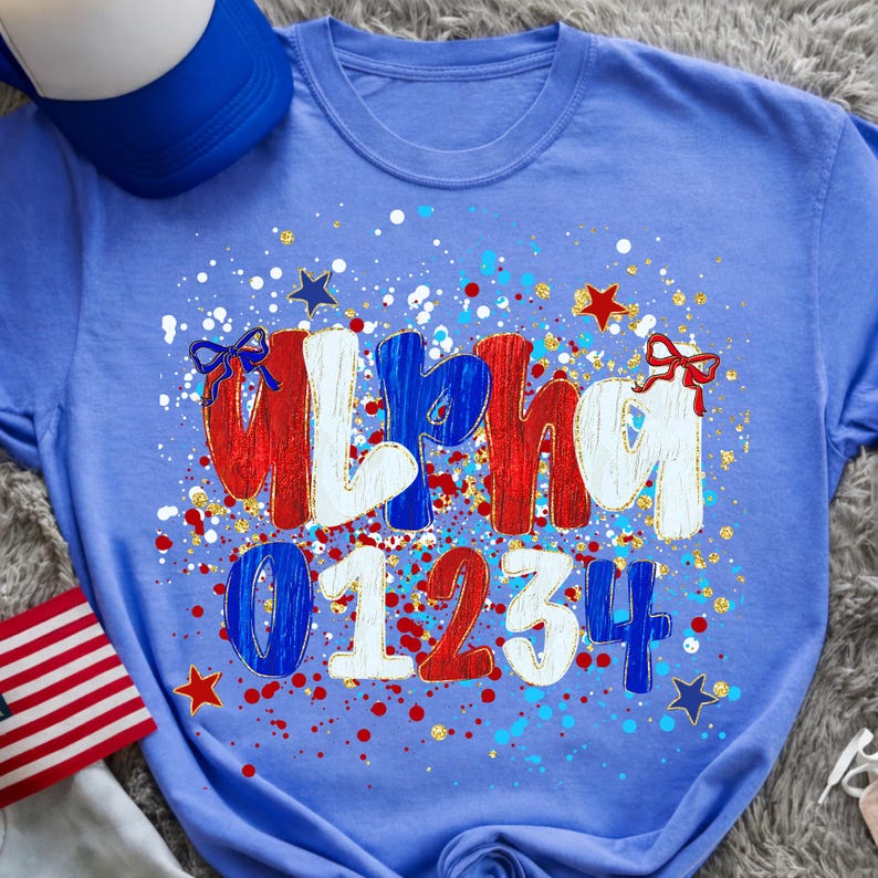 May include: A light blue t-shirt with the word "Alpha" in red, white, and blue, and the numbers "01234". The design includes stars, bows, and paint splatters in red, white, blue, and gold. A blue and white trucker hat is visible.