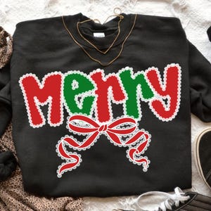 May include: Black sweatshirt with the word "MERRY" in red and green, outlined in white lace. A red bow with white lace detailing is below the word. A gold necklace is draped over the sweatshirt.
