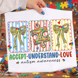 May include: White t-shirt with a colourful design promoting autism awareness. The design includes three framed sections with a bow, puzzle piece, and bow. The words "ACCEPT-UNDERSTAND-LOVE" and "autism awareness" are printed on the shirt.