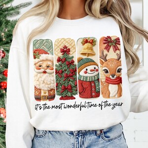 May include: White sweatshirt featuring four Christmas-themed designs: Santa Claus, a Christmas tree, a snowman, and a reindeer. The text "It's the most wonderful time of the year" is printed below the designs. The sweatshirt is worn by a person.
