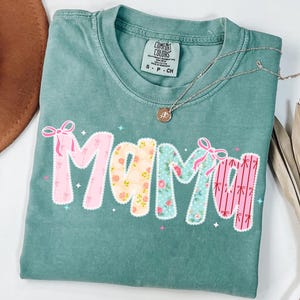 May include: A teal Comfort Colors t-shirt with the word "Mama" in a patchwork design. The letters are decorated with floral patterns and pink bows. A silver necklace with a small pendant rests on the shirt.