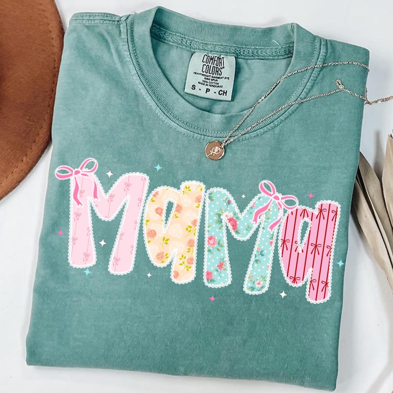 May include: A teal Comfort Colors t-shirt with the word "Mama" in a patchwork design. The letters are decorated with floral patterns and pink bows. A silver necklace with a small pendant rests on the shirt.