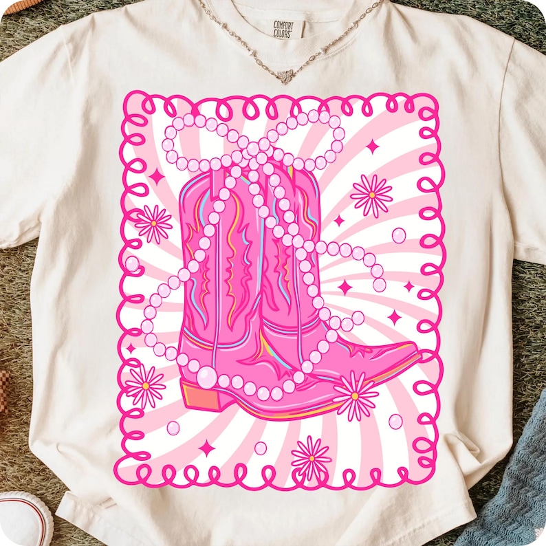 May include: Cream-colored t-shirt featuring a pink graphic of cowboy boots adorned with a pearl necklace. The design includes a pink border, flowers, and a striped background. The shirt has a Comfort Colors tag.