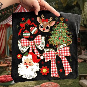 May include: Black t-shirt with festive Christmas graphics. The design includes a reindeer with a red bow, a decorated Christmas tree, Santa Claus, red and white checkered bows, baubles, and red stars and flowers.