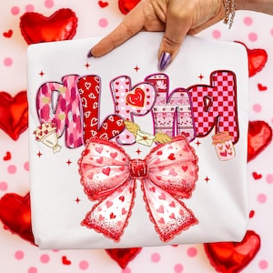 May include: White sweatshirt with the word "ALHAM" in a colorful, Valentine's Day-themed design. The letters feature hearts, patterns, and a large red bow with heart details. The background includes red heart-shaped balloons.