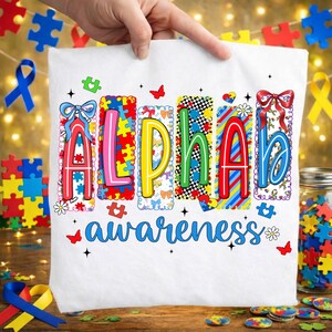 May include: White t-shirt featuring the word "ALPHA" in vibrant puzzle-piece letters, and the word "awareness" in blue script. The shirt is held up by a hand, with puzzle pieces and ribbons in the background.