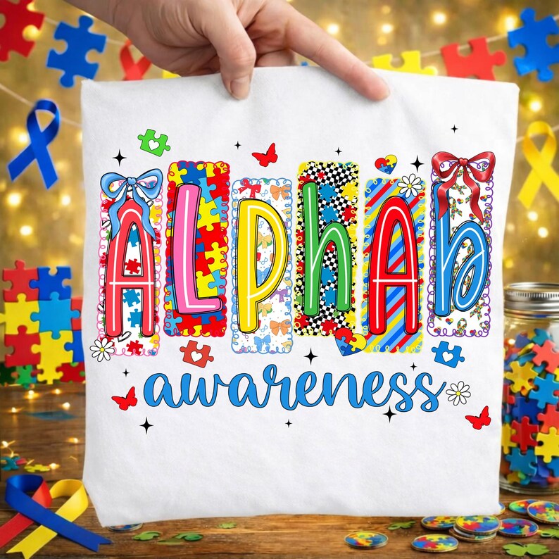 May include: White t-shirt featuring the word "ALPHA" in vibrant puzzle-piece letters, and the word "awareness" in blue script. The shirt is held up by a hand, with puzzle pieces and ribbons in the background.