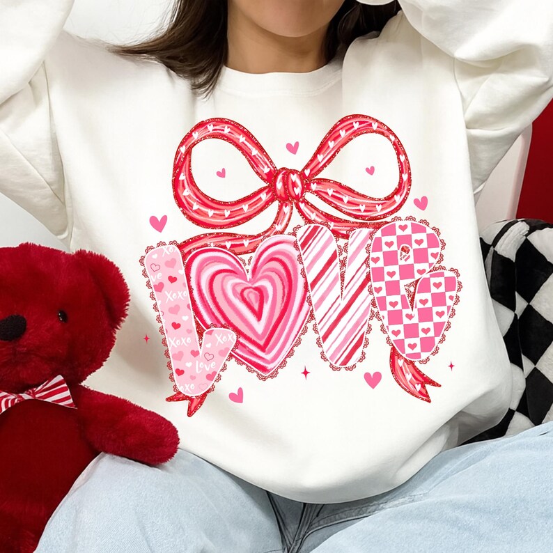 May include: White sweatshirt with a large red bow and the word "LOVE" in pink, featuring heart and candy cane designs. Small pink hearts are scattered around the design. A red teddy bear is visible on the left.