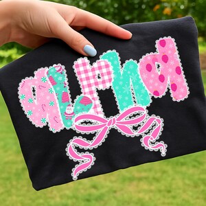 Pu&ograve; includere: T-shirt nera con la scritta "MAMA" in rosa, verde e motivi a scacchi, decorata con fiocchi di neve, alberi di Natale e ornamenti. Un fiocco rosa con bordo in pizzo bianco &egrave; in basso.