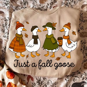 May include: Beige sweatshirt with a graphic of four cartoon geese dressed in fall attire. The geese wear hats, scarves, and coats, with one holding a coffee cup. The text "Just a fall goose" is printed below the geese. Fall-themed apparel.