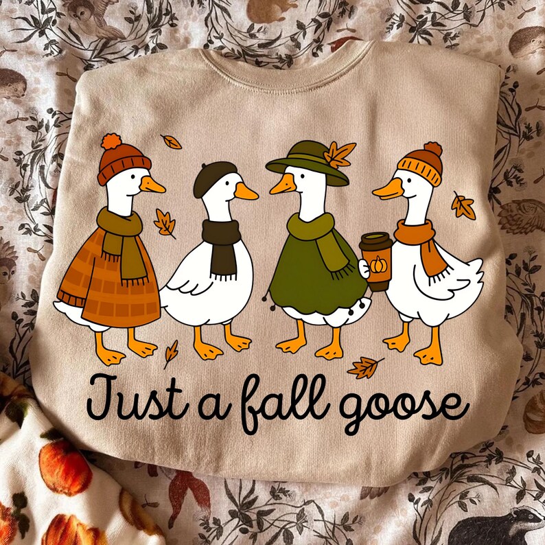 May include: Beige sweatshirt with a graphic of four cartoon geese dressed in fall attire. The geese wear hats, scarves, and coats, with one holding a coffee cup. The text "Just a fall goose" is printed below the geese. Fall-themed apparel.